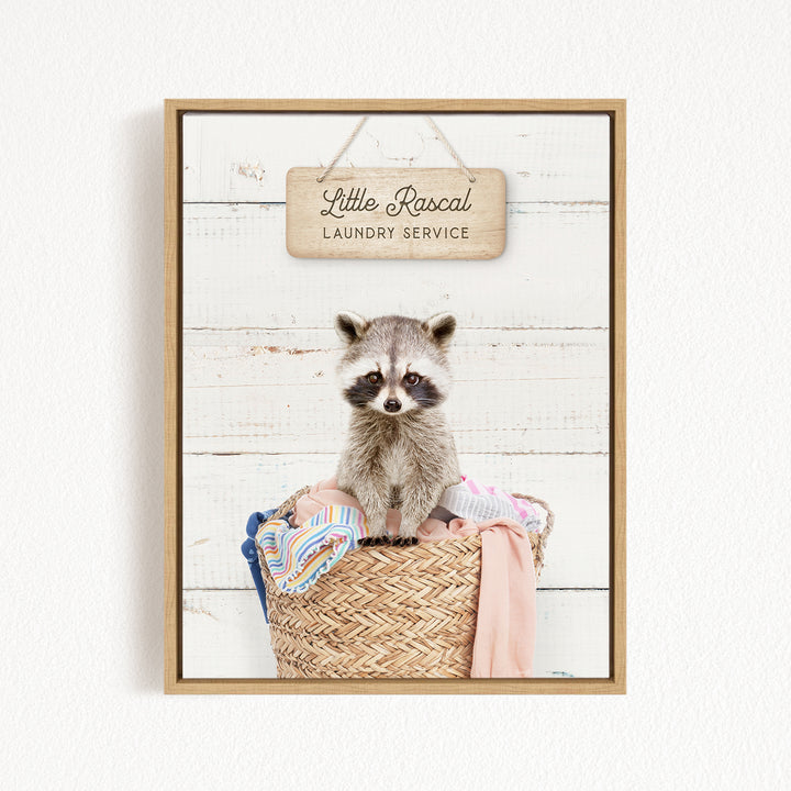 A raccoon sitting in a woven laundry basket, with a sign above that reads "Little Racal Laundry Service".