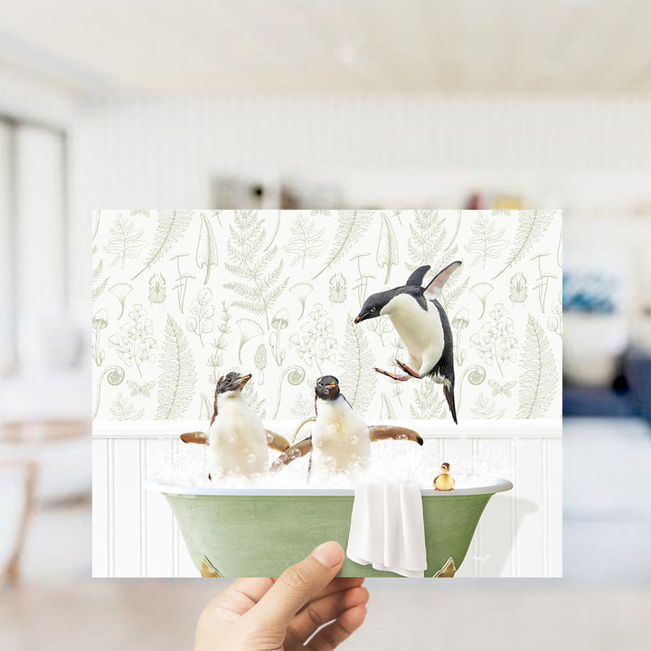 A hand holds up a framed image depicting three penguins sitting in a bathtub, with one penguin standing on the edge of the tub.
