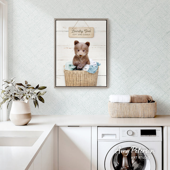 a framed picture of a brown bear sitting in a wicker basket, placed on a white countertop in a laundry room.
