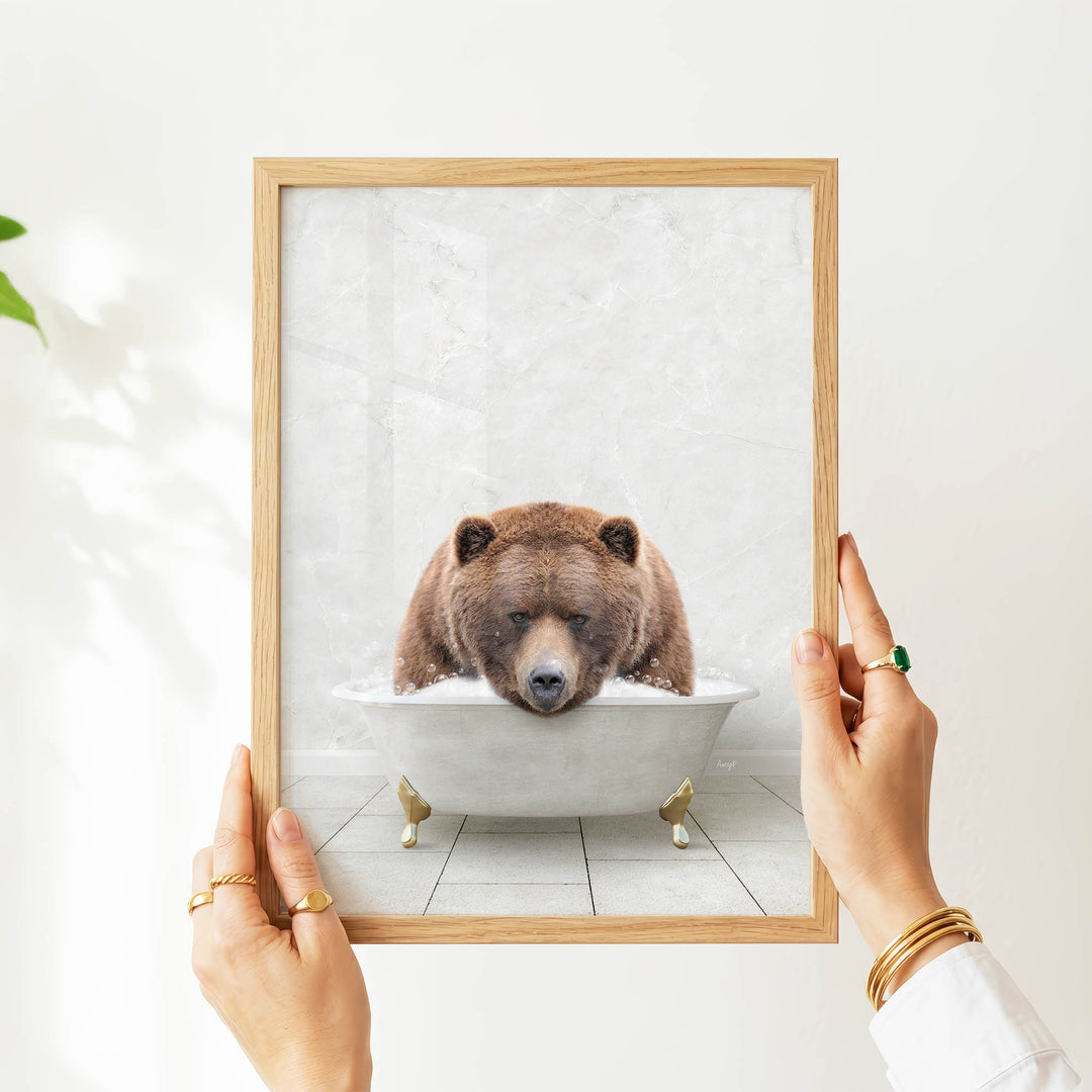 A person's hands are holding a framed picture of a brown bear sitting in a bathtub.