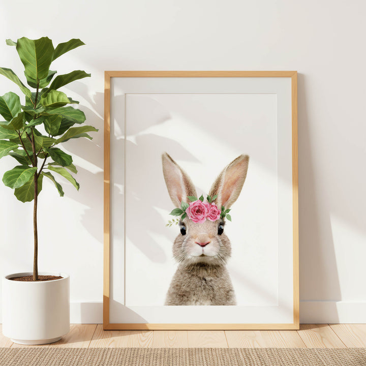 A framed picture of a rabbit wearing a pink flower crown is displayed on a white wall, with a potted plant to the left.