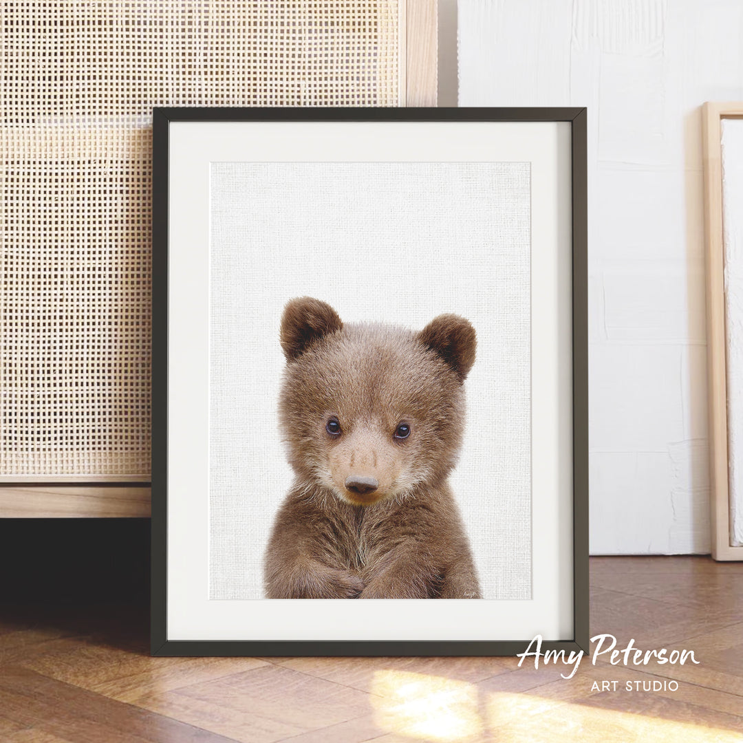 A framed photograph of a cute brown bear cub is displayed on a wooden floor in front of a woven basket.