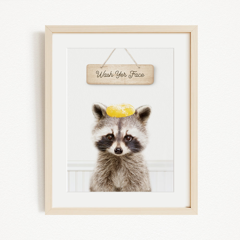A raccoon wearing a yellow hat and looking directly at the camera, with a wooden sign above that says "Wash Your Face".