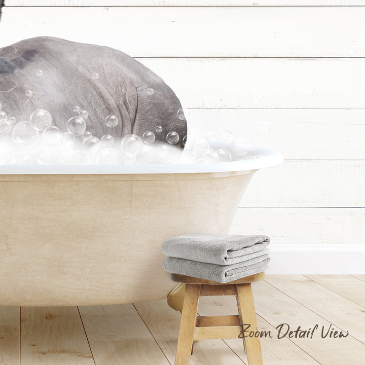 A large bathtub filled with bubbles, with a gray elephant partially submerged in the water. A wooden stool is placed next to the tub, and there are two gray towels nearby.
