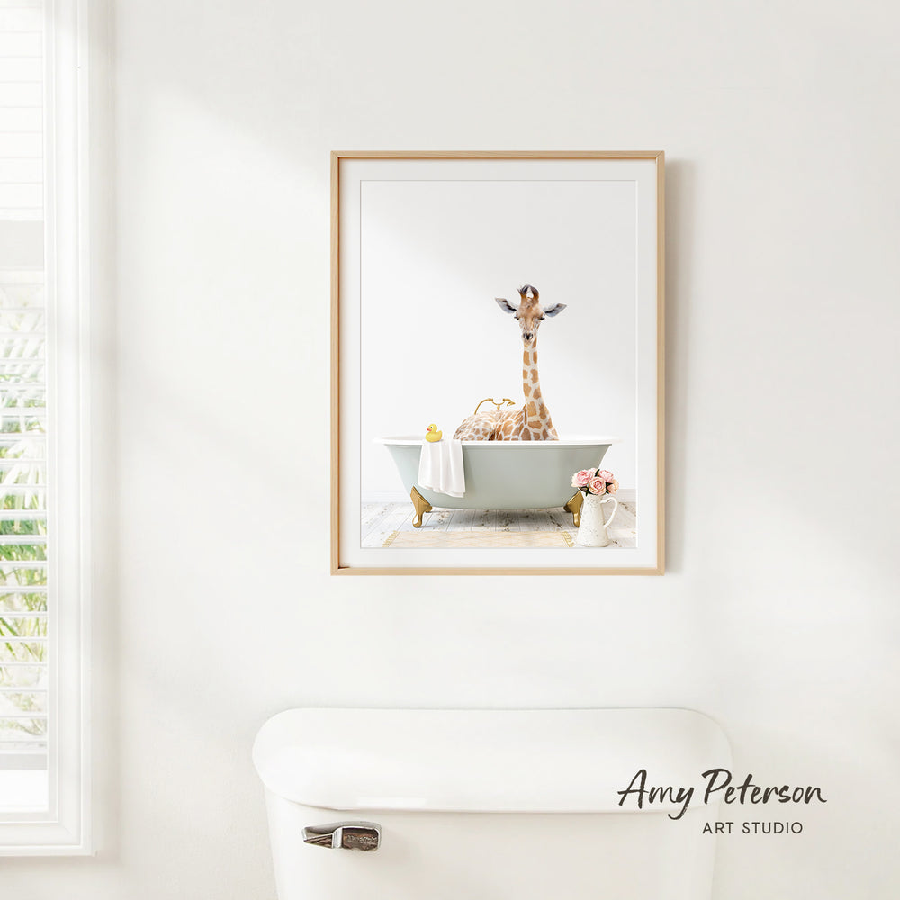 A framed image of a giraffe sitting in a bathtub, surrounded by various objects such as a vase and a towel, is displayed on a white wall in a bathroom.