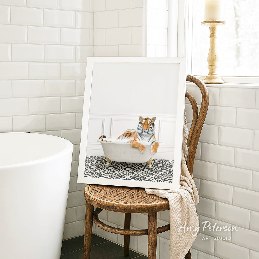A framed image of a tiger sitting in a bathtub, with a towel draped over the back of a chair in a bathroom setting.