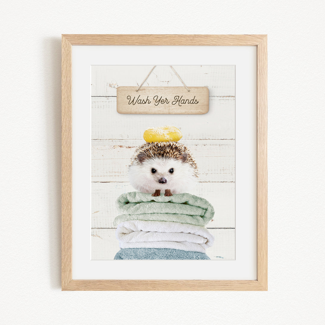 A framed image of a hedgehog sitting on a stack of towels, with a wooden sign above it that says "Wash Your Hands".