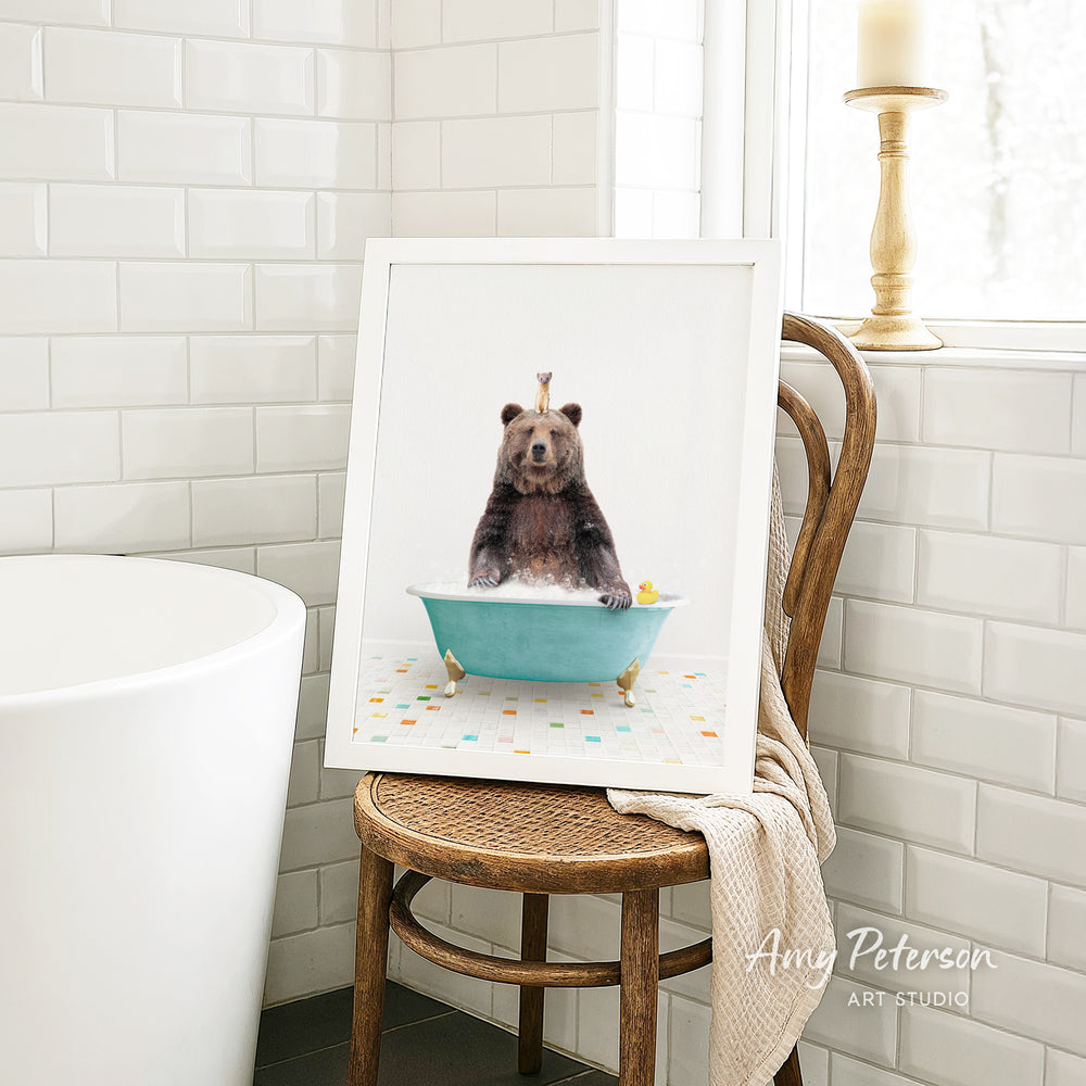 A framed image of a bear sitting in a bathtub, surrounded by bath toys, is displayed on a wooden chair in a bathroom setting.