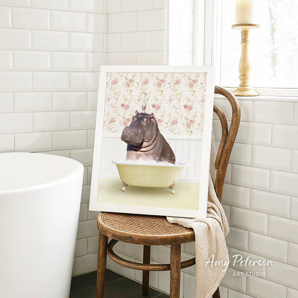 A framed image of a hippopotamus sitting in a bathtub is displayed on a wooden chair in a bathroom setting.
