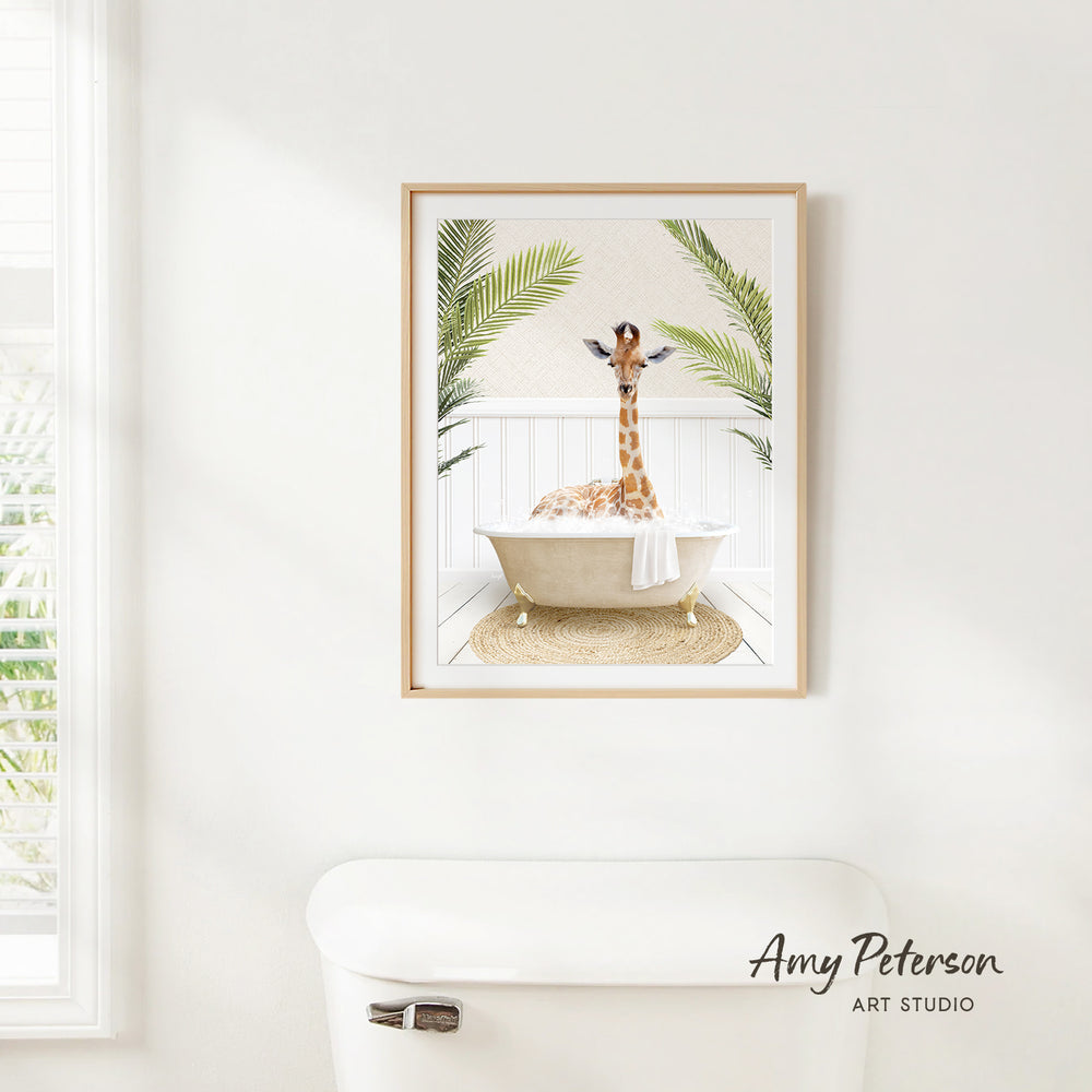A framed print of a giraffe sitting in a bathtub, surrounded by palm leaves, hangs on a white wall.