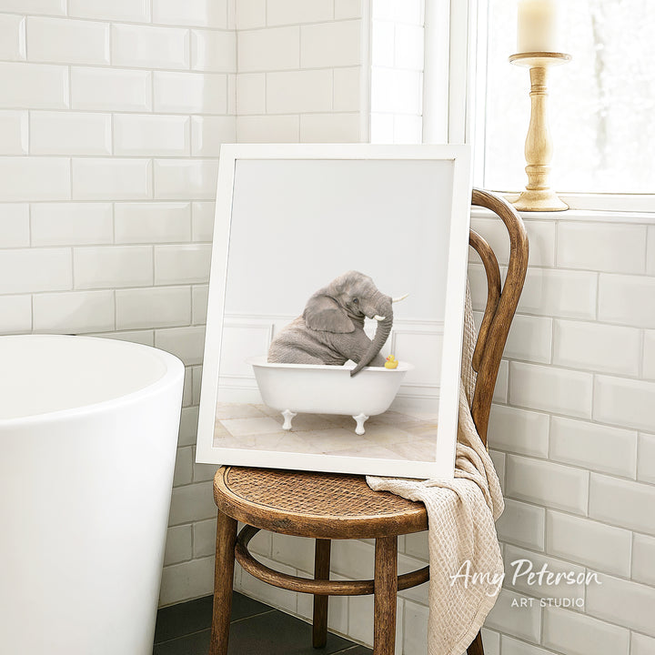A framed photograph of a gray elephant sitting in a white bathtub, with a towel draped over the back of a wooden chair in a bathroom setting.