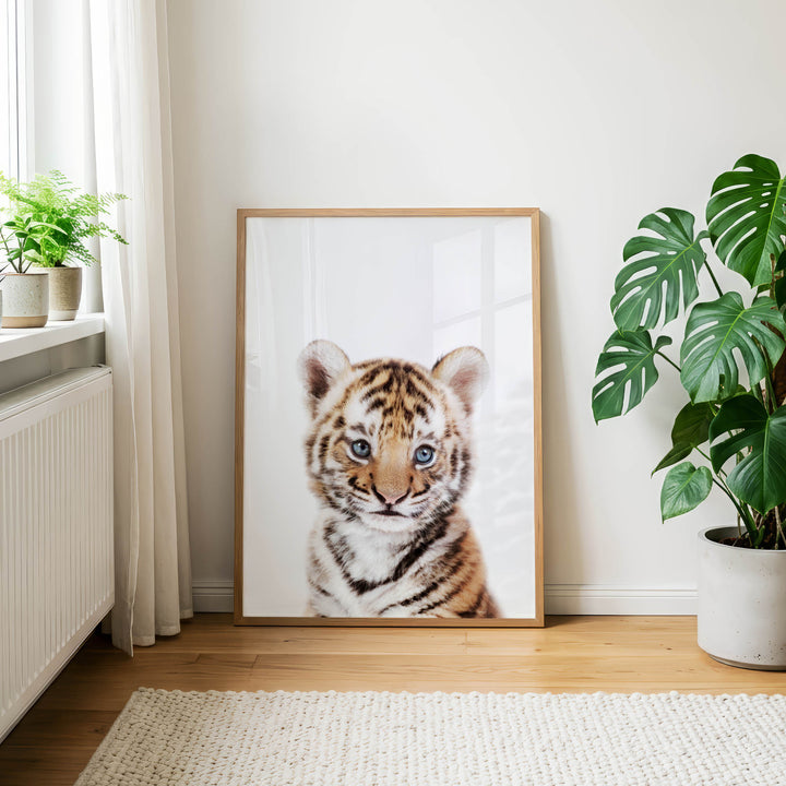a large framed picture of a tiger cub, which is the main focus of the scene. The tiger cub is positioned in the center of the frame, surrounded by a white background. The room has a wooden floor and a white rug, creating a cozy and natural atmosphere. There are also two potted plants in the room, one on the left side and the other on the right side, adding a touch of greenery to the space.