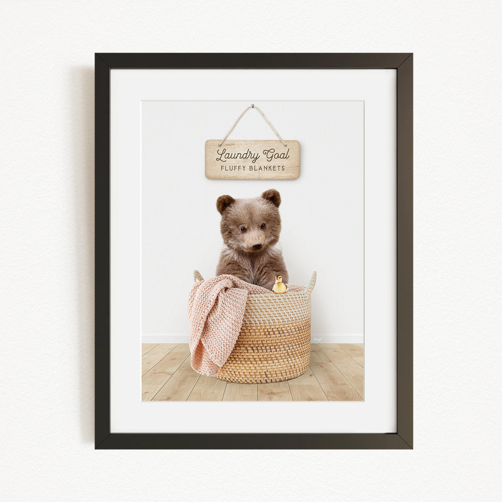 A framed image of a brown bear sitting in a woven basket, with a sign above it that says "Laundry Goal Fluffy Blankets".
