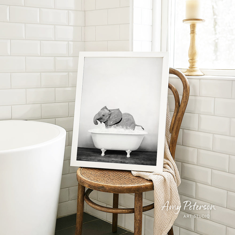 A framed black and white photograph of an elephant sitting in a bathtub, placed on a wooden chair in a bathroom setting.