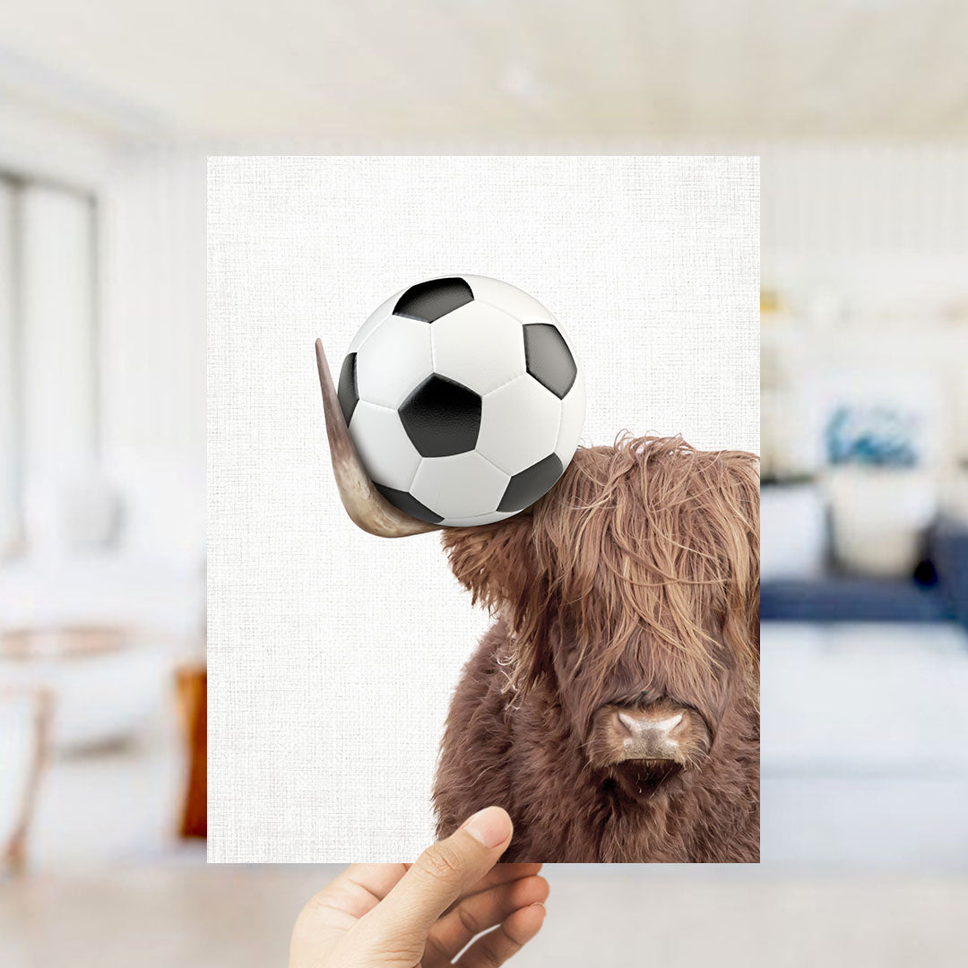 A hand holds a picture of a brown cow with a soccer ball on its head, creating a playful and whimsical scene.