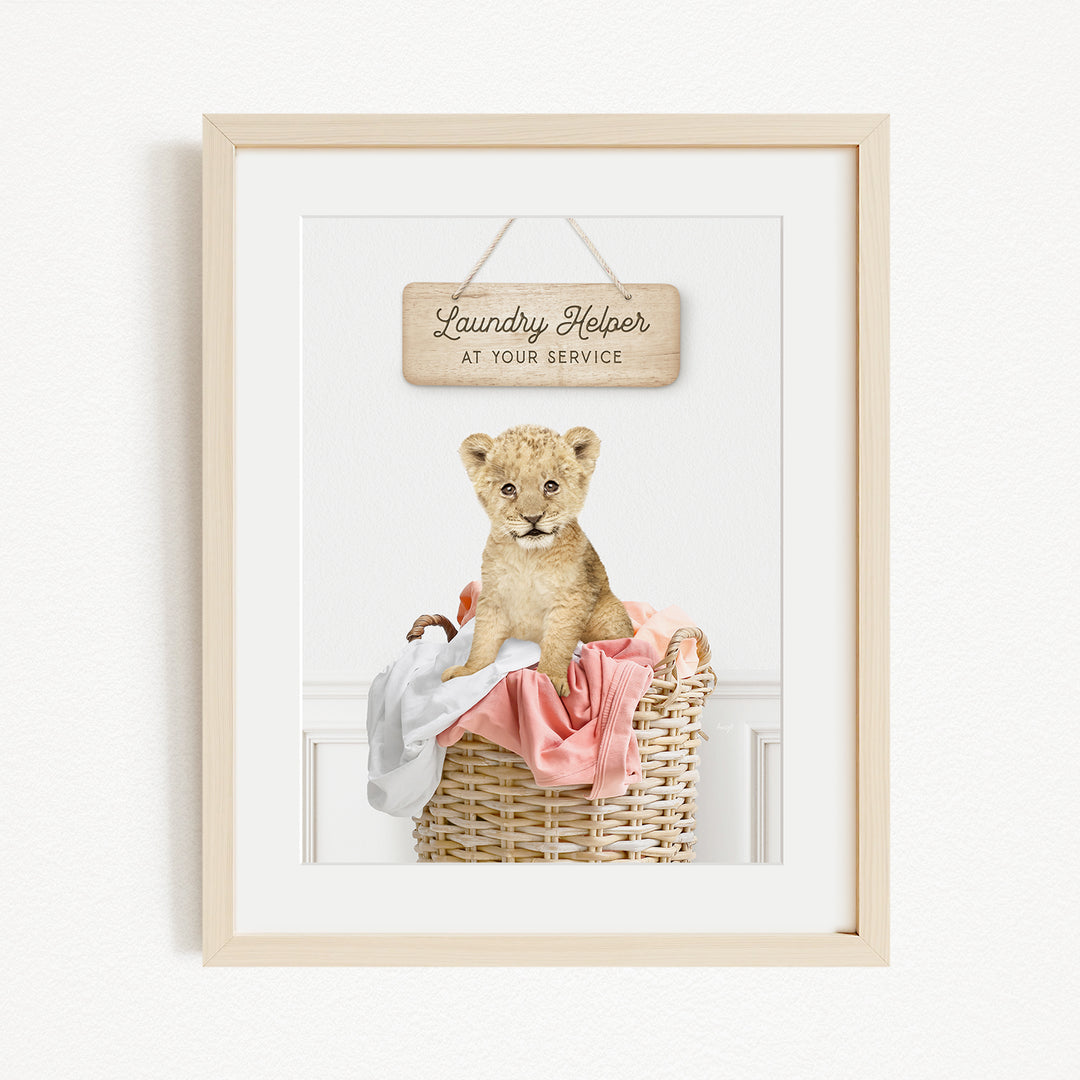 A framed image of a young lion cub sitting in a wicker basket filled with clothes, with a sign above that reads "Laundry Helper at your service".
