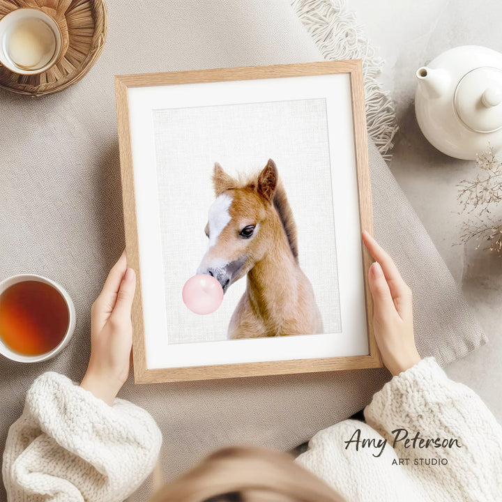 A person is holding a framed picture of a horse blowing a bubble with a pink bubble gum bubble.
