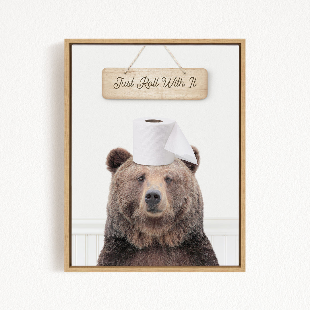 A framed image of a brown bear wearing a roll of toilet paper on its head, with a wooden sign above it that says "Just Roll With It".