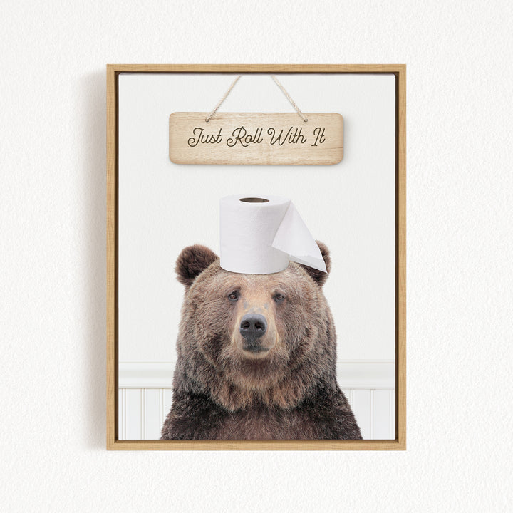 A framed image of a brown bear wearing a roll of toilet paper on its head, with a wooden sign above it that says "Just Roll With It".