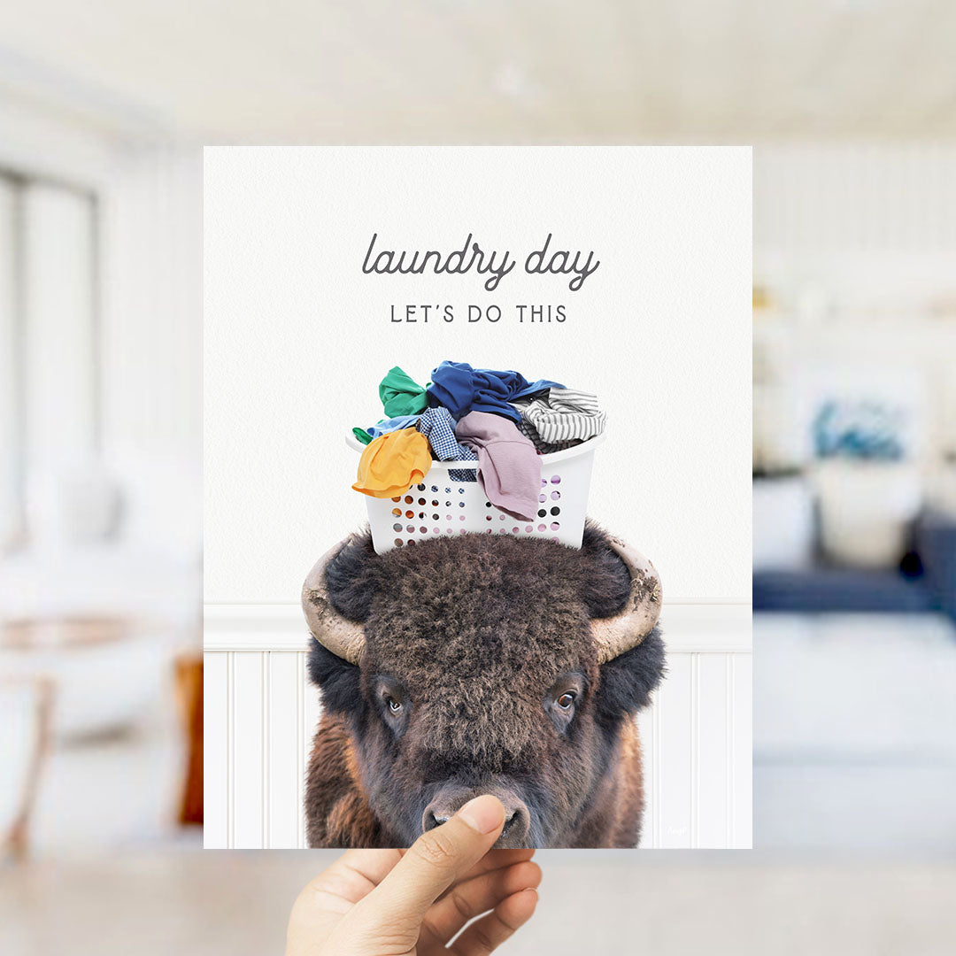 A hand holding a poster with a bison wearing a laundry basket on its head, with the text "laundry day let's do this" written above it.