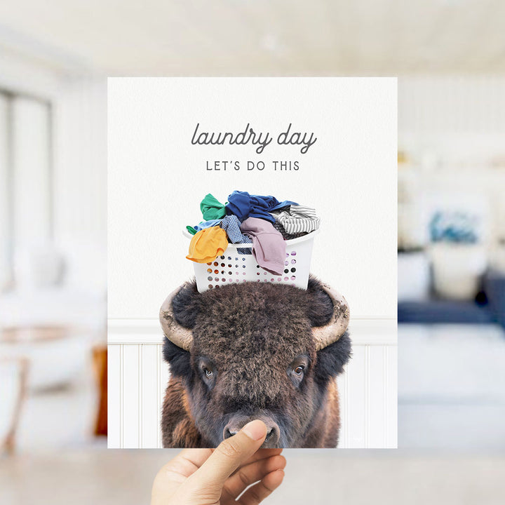 A hand holding a poster with a bison wearing a laundry basket on its head, with the text "laundry day let's do this" written above it.