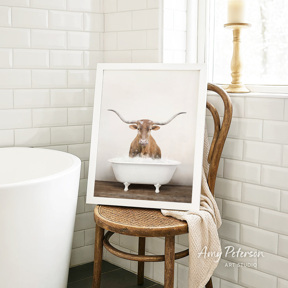 A framed image of a bull with long horns sitting in a white bathtub is displayed on a wooden chair in a bathroom.