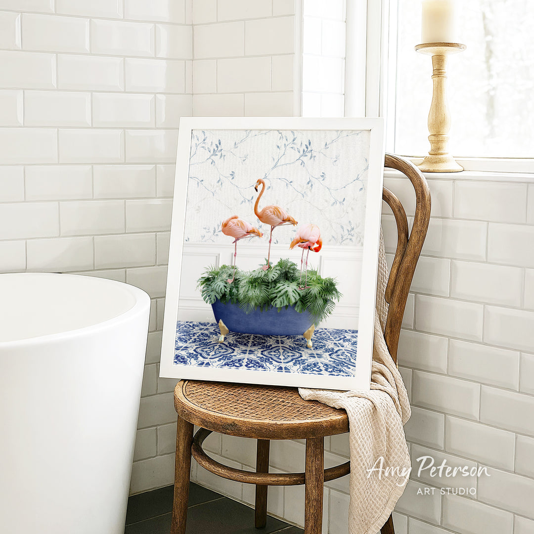 A framed image of three pink flamingos standing in a blue and white patterned vase, placed on a wooden chair in a bathroom setting.