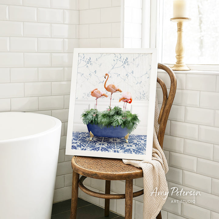 A framed image of three pink flamingos standing in a blue and white patterned vase, placed on a wooden chair in a bathroom setting.