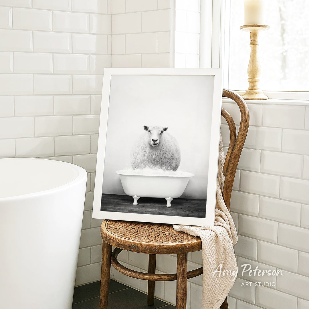 A framed black and white photograph of a sheep sitting in a bathtub is displayed on a wooden chair in a bathroom setting.