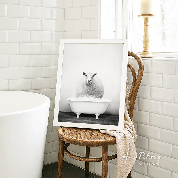 A framed black and white photograph of a sheep sitting in a bathtub is displayed on a wooden chair in a bathroom setting.
