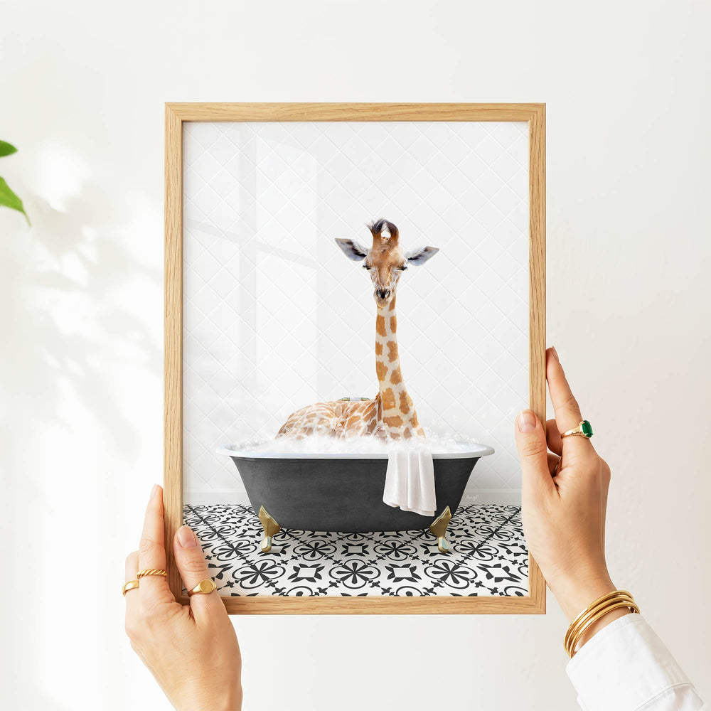 A person's hands are holding a framed picture of a giraffe sitting in a bathtub filled with water and bubbles.
