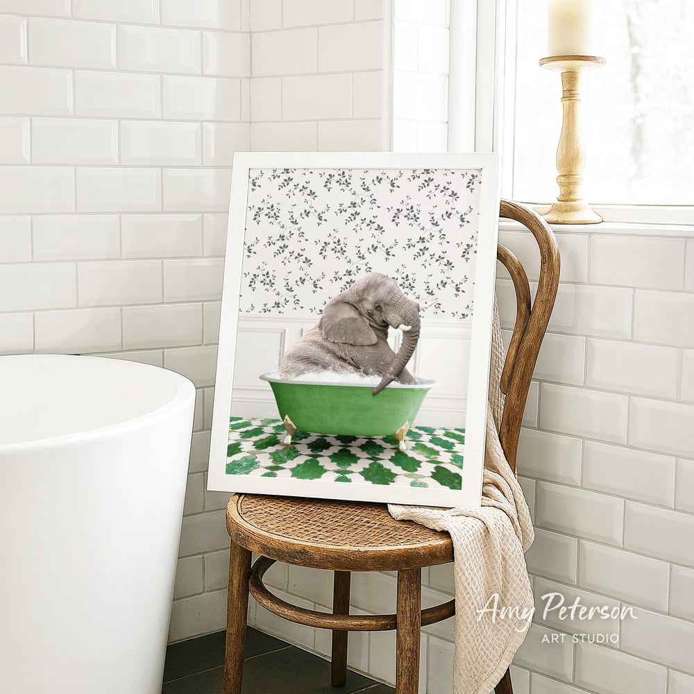 A framed image of a gray elephant sitting in a green bathtub, surrounded by a floral pattern, is displayed on a wooden chair in a bathroom setting.