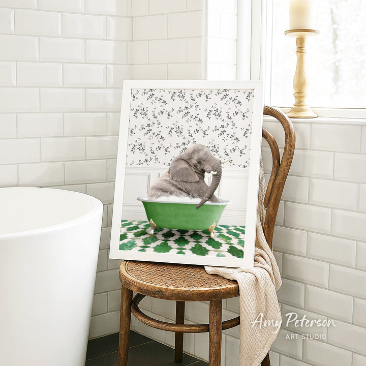 A framed image of a gray elephant sitting in a green bathtub, surrounded by a floral pattern, is displayed on a wooden chair in a bathroom setting.