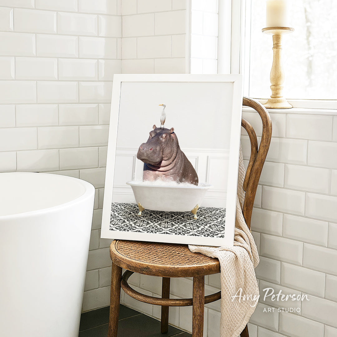 A framed image of a hippopotamus sitting in a bathtub, with a bird perched on its head, is displayed on a wooden chair in a bathroom setting.