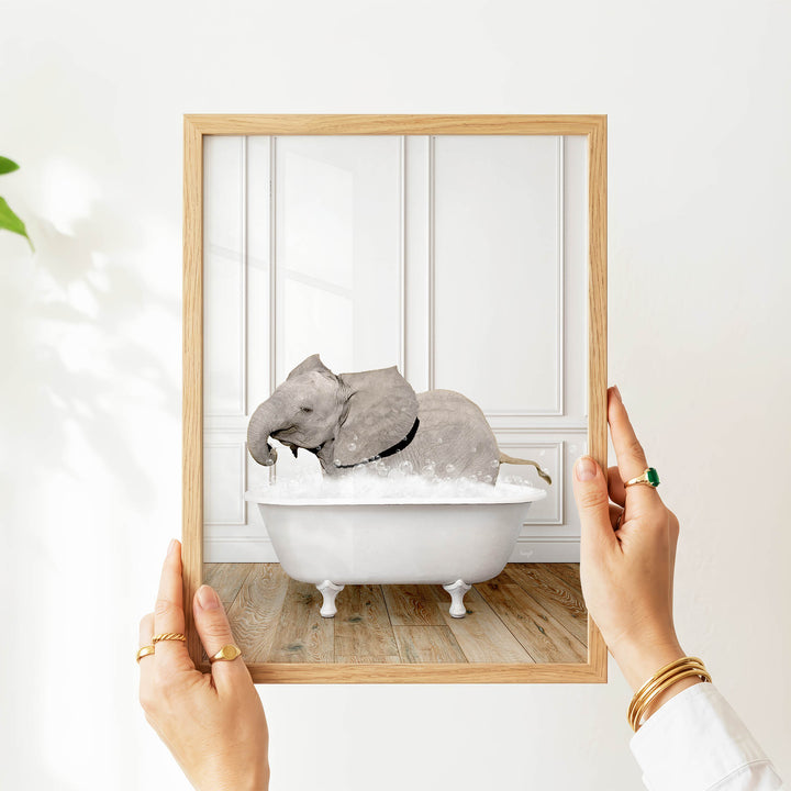A person's hands are holding a picture frame with a photograph of a gray elephant sitting in a white bathtub filled with water.
