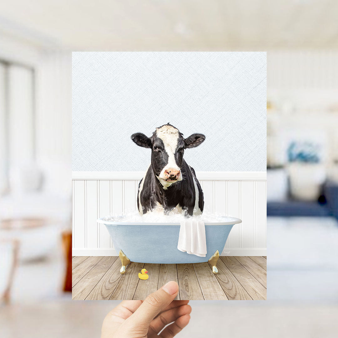 A black and white cow is sitting in a blue bathtub filled with water, with a yellow rubber duck floating nearby.