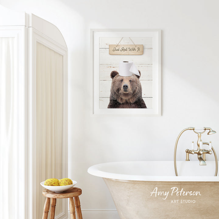 a bathroom with a framed picture of a bear wearing a toilet paper hat, a wooden stool with a bowl of fruit, and a bathtub with a faucet.
