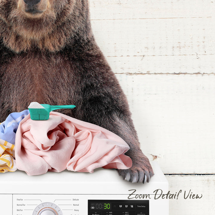 A brown bear is sitting on top of a washing machine, with laundry items such as clothes and a detergent bottle nearby.