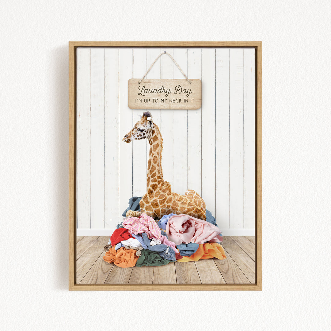 A giraffe is sitting on a pile of clothes in front of a wooden sign that says "Laundry Day" and "I'm up to my neck in it".