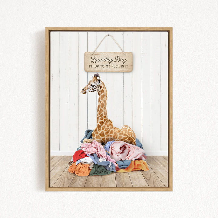 A giraffe is sitting on a pile of clothes in front of a wooden sign that says "Laundry Day" and "I'm up to my neck in it".
