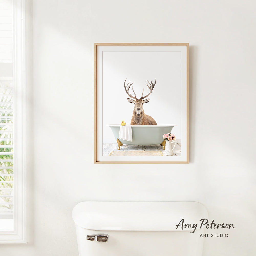 A framed image of a deer sitting in a bathtub, with a vase of flowers and a white toilet in the foreground.