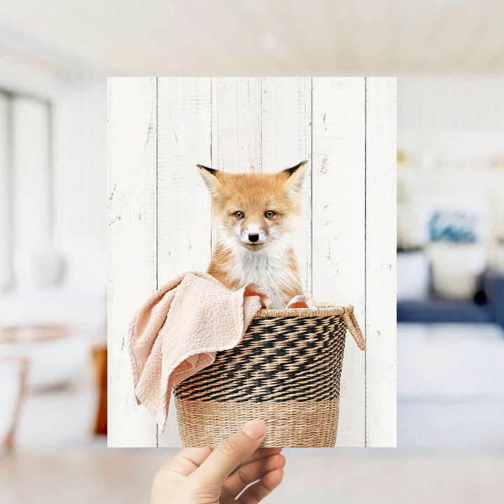 A small orange fox is sitting in a woven basket, with a pink towel draped over it.