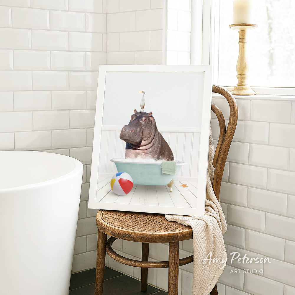 A framed image of a hippopotamus sitting in a bathtub, surrounded by a beach ball, a towel, and a bird, is displayed on a wooden chair in a bathroom setting.