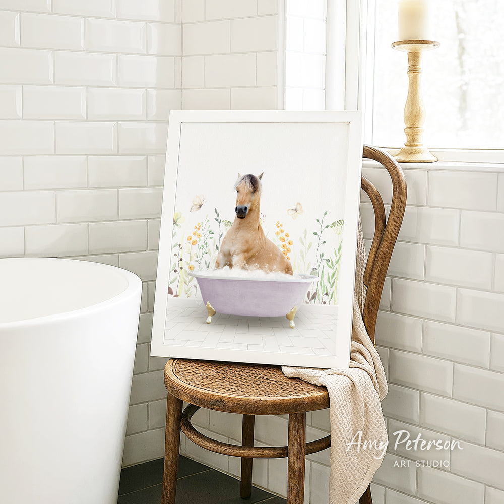 A framed image of a horse sitting in a bathtub, surrounded by flowers and butterflies, is displayed on a wooden chair in a bathroom setting.