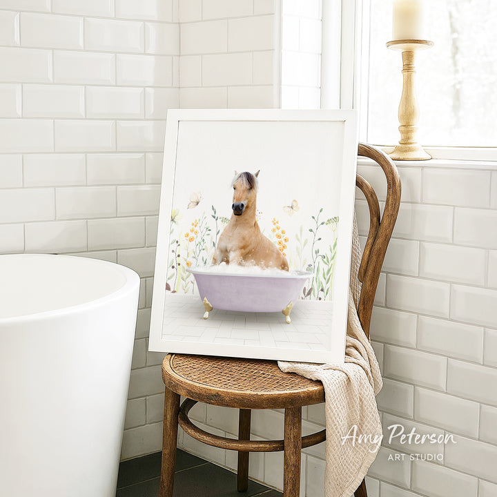 A framed image of a horse sitting in a bathtub, surrounded by flowers and butterflies, is displayed on a wooden chair in a bathroom setting.