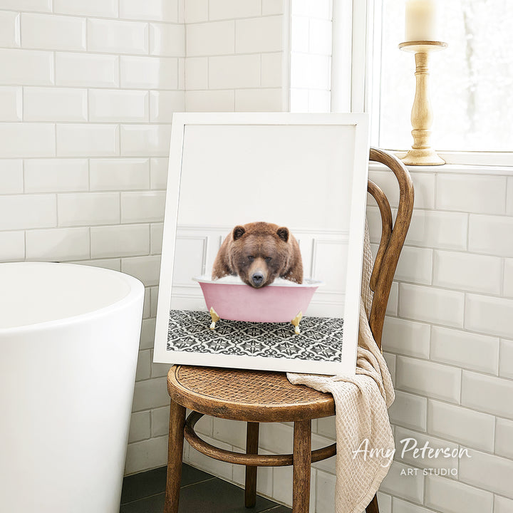A framed image of a bear sitting in a pink bathtub, with a towel draped over the back of a wooden chair in a bathroom setting.