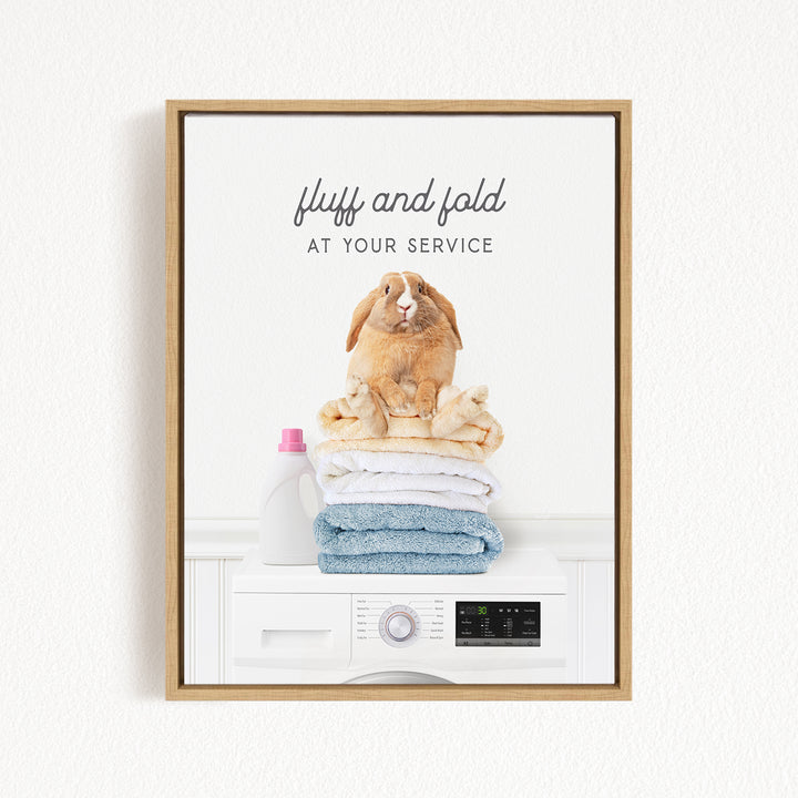 A framed image of a rabbit sitting on top of a stack of folded towels, with the text "fluff and fold at your service" above the rabbit.