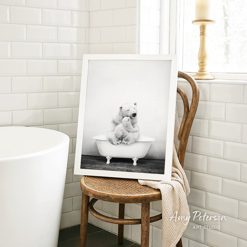 A framed black and white photograph of two polar bears sitting in a bathtub is displayed on a wooden chair in a bathroom setting.