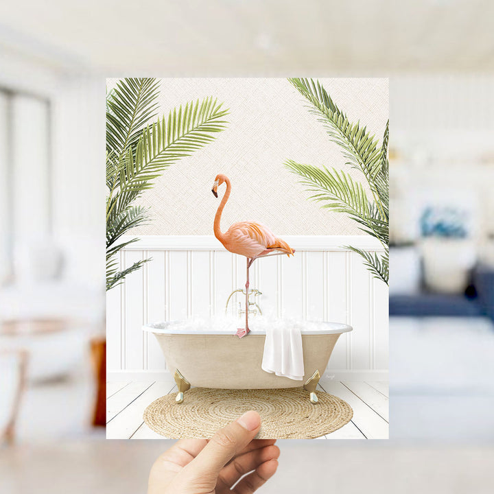 A hand holds up a framed image of a pink flamingo standing on a white bathtub, surrounded by green palm leaves.