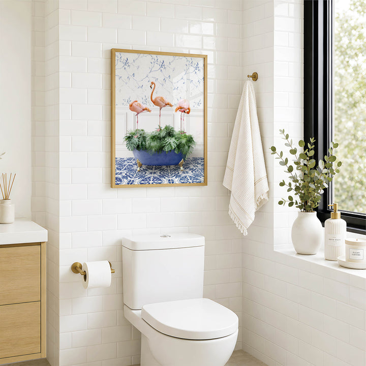 a bathroom with a white toilet, a framed picture of three flamingos in a blue and white vase, and a window with a plant on the sill.
